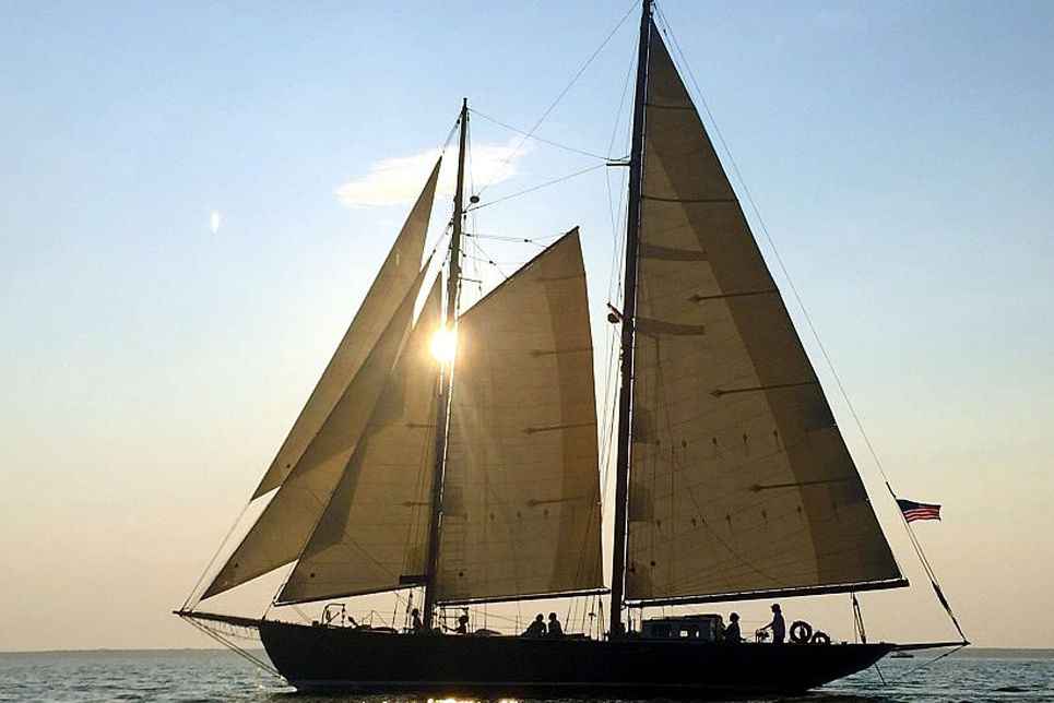 A tall ship with sails raised glides across the water, silhouetted against a sunset sky. An American flag flutters at the stern.